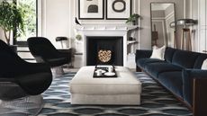 Modern living room with dark blue sofa and armchairs, padded cream coffee table, patterned rug, large mirror and fireplace with neatly arranged logs.