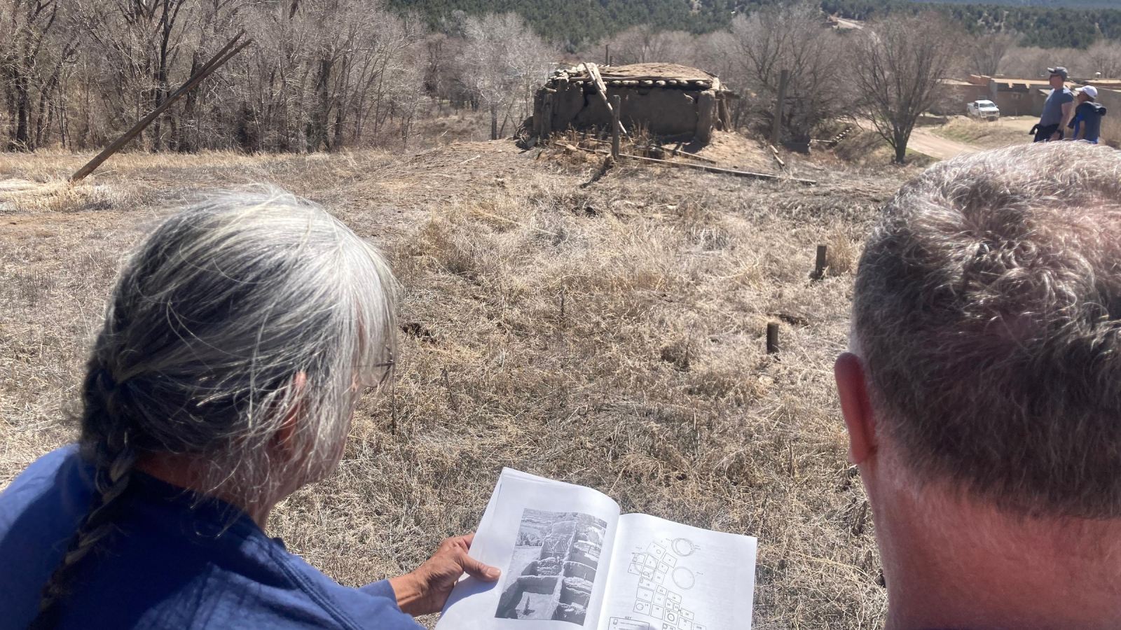 Two people stand by an ancient site in a field looking through a document.