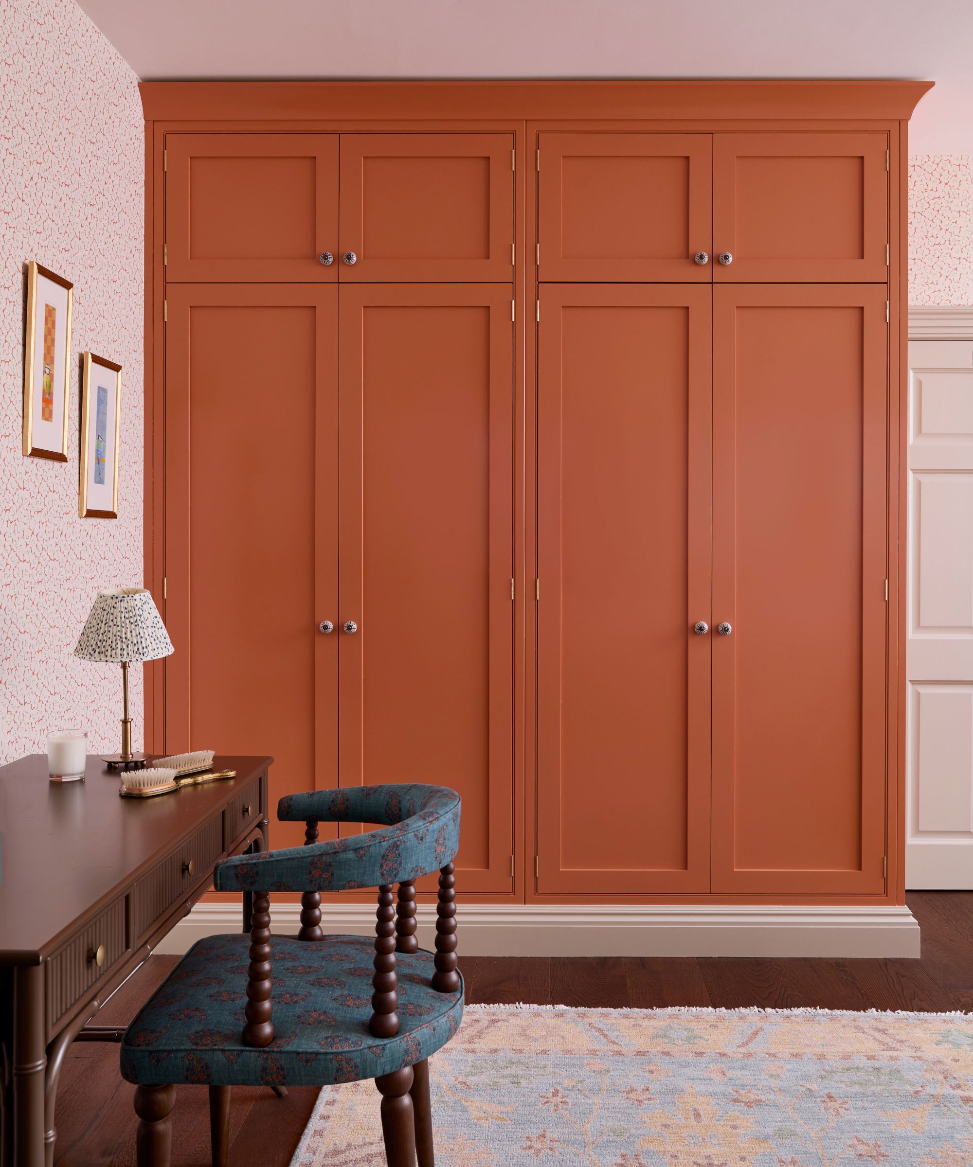 Bedroom with pale pink wallpapered walls, a mahogany desk and bobbin chair, and built-in wardrobes painted deep orange, with wooden floors and a blue, cream and orange Persian style rug