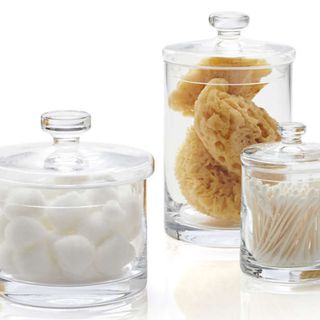Three clear glass jars with lids each storing different items found in bathrooms such as Q-tips sponges and cotton wool balls