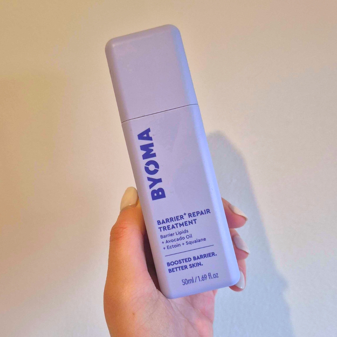 Hand holding the purple BYOMA Barrier+ Repair Treatment against a white wall