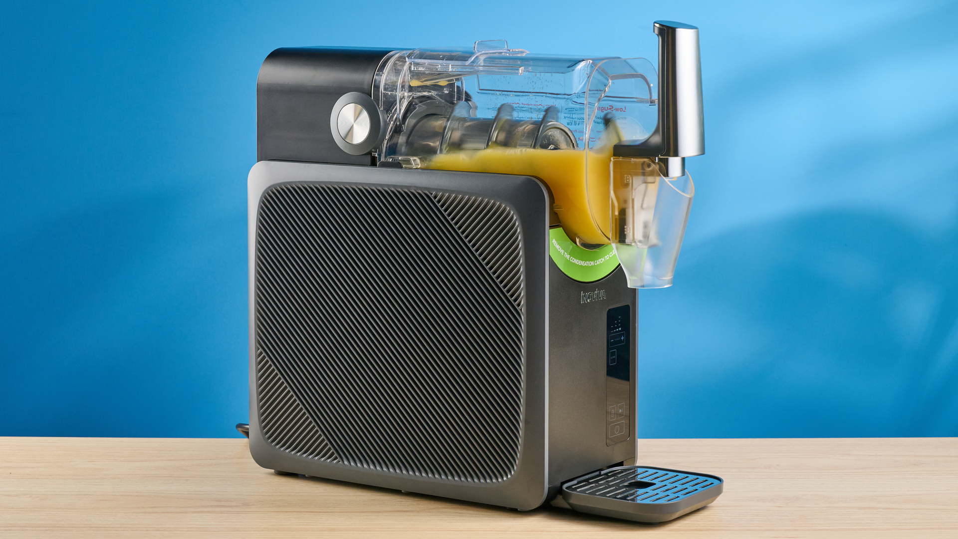 the inoviva slushie machine, a slushie maker with a plastic cylinder body, a dispensing spout, photographed making orange juice slush against a blue tom&#039;s guide background