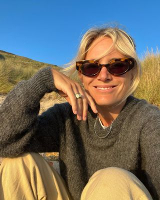 Woman wearing sunglasses, gray sweater, and khaki pants holding up hand to camera with chunky gold engagement ring.