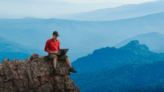 A man using a laptop sitting atop a rocky outcrop against a backdrop of mountains.