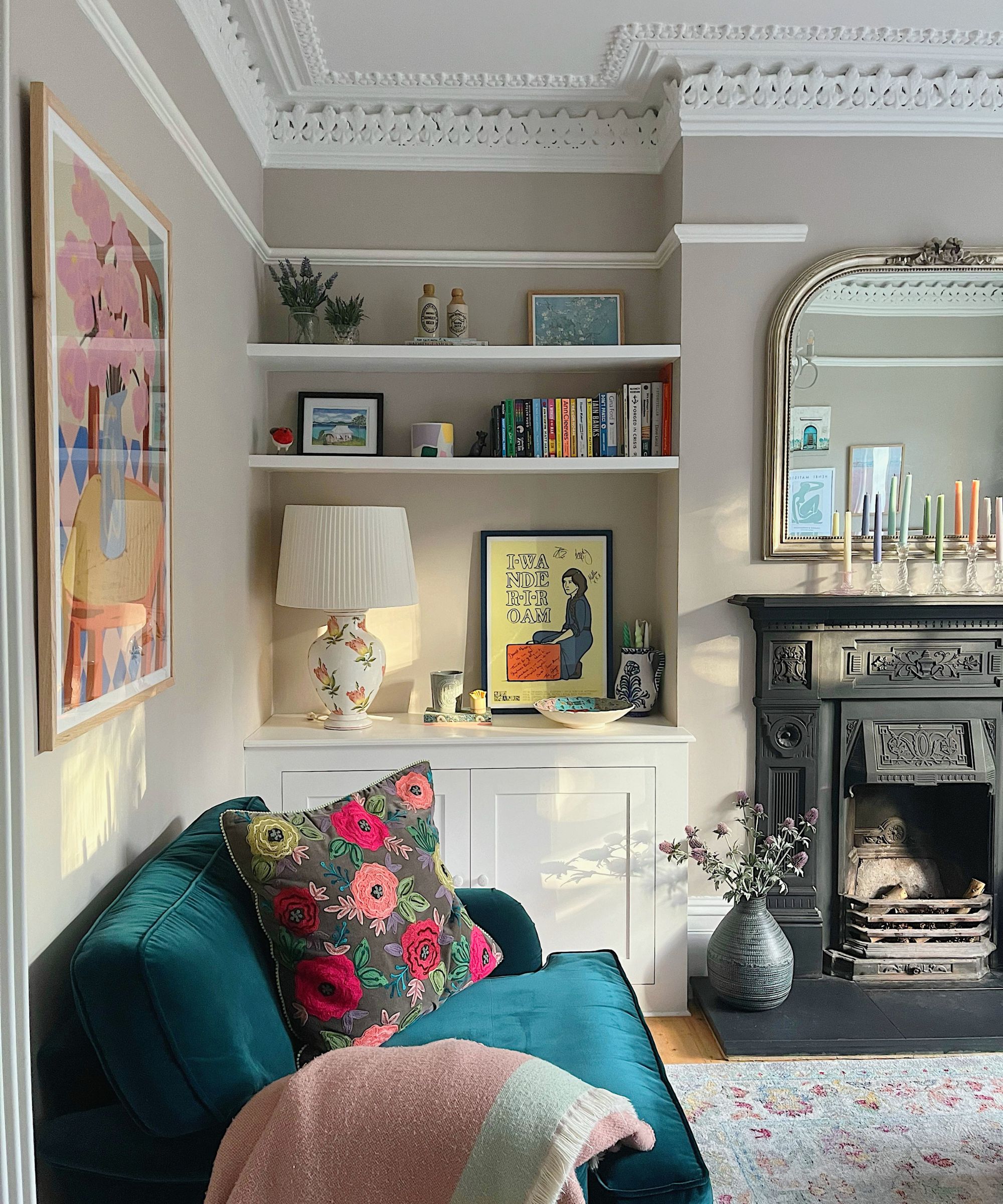 Living room with crown moulding, Farrow &amp;amp; Ball's Elephant Breath on the walls, a built-in alcove with bookshelves and artwork, a blue velvet sofa, a Persian rug and a large Victorian black fireplace.