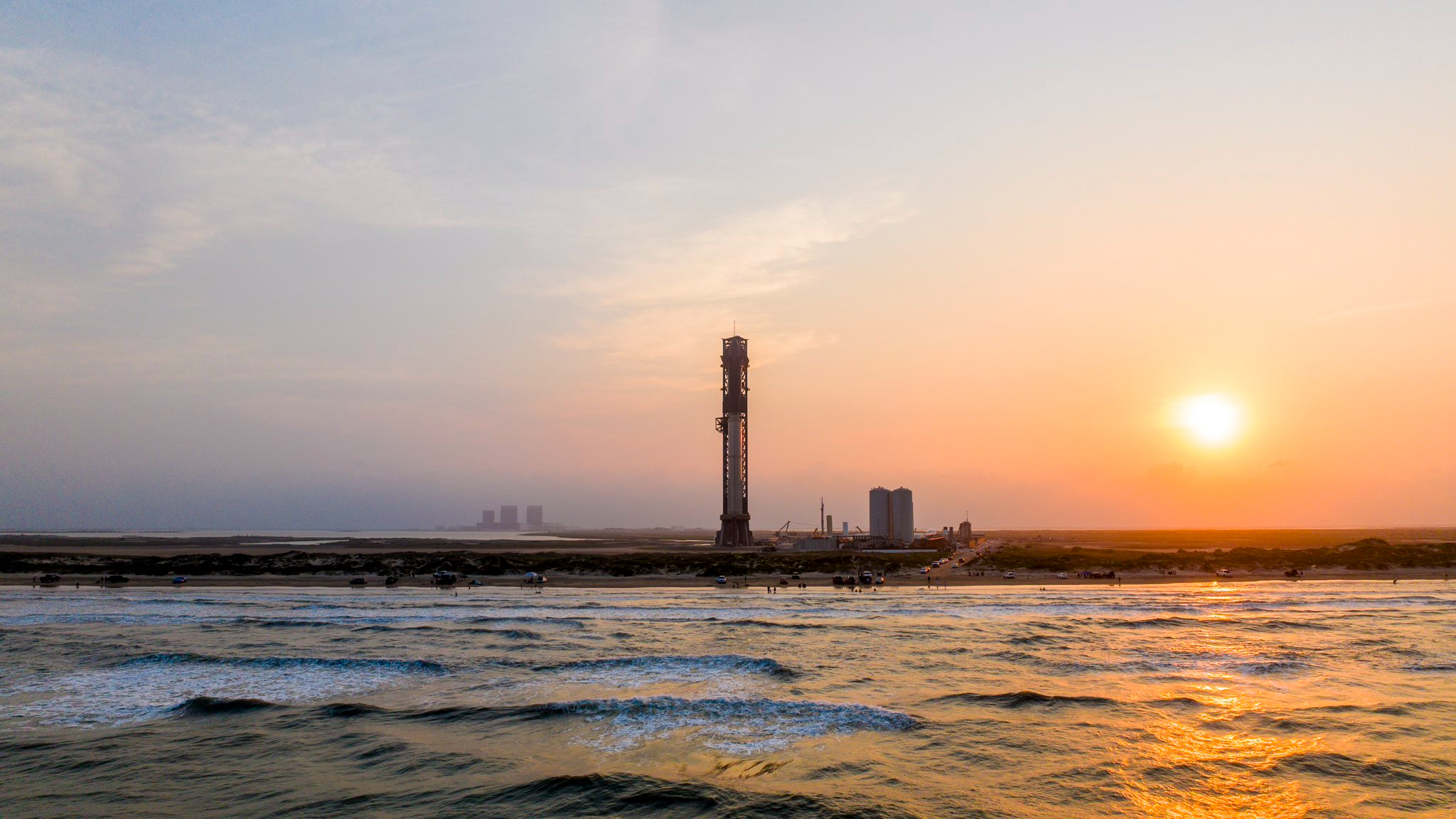 view of a giant rocket on the launch pad with the ocean in the foreground and the setting sun in the background