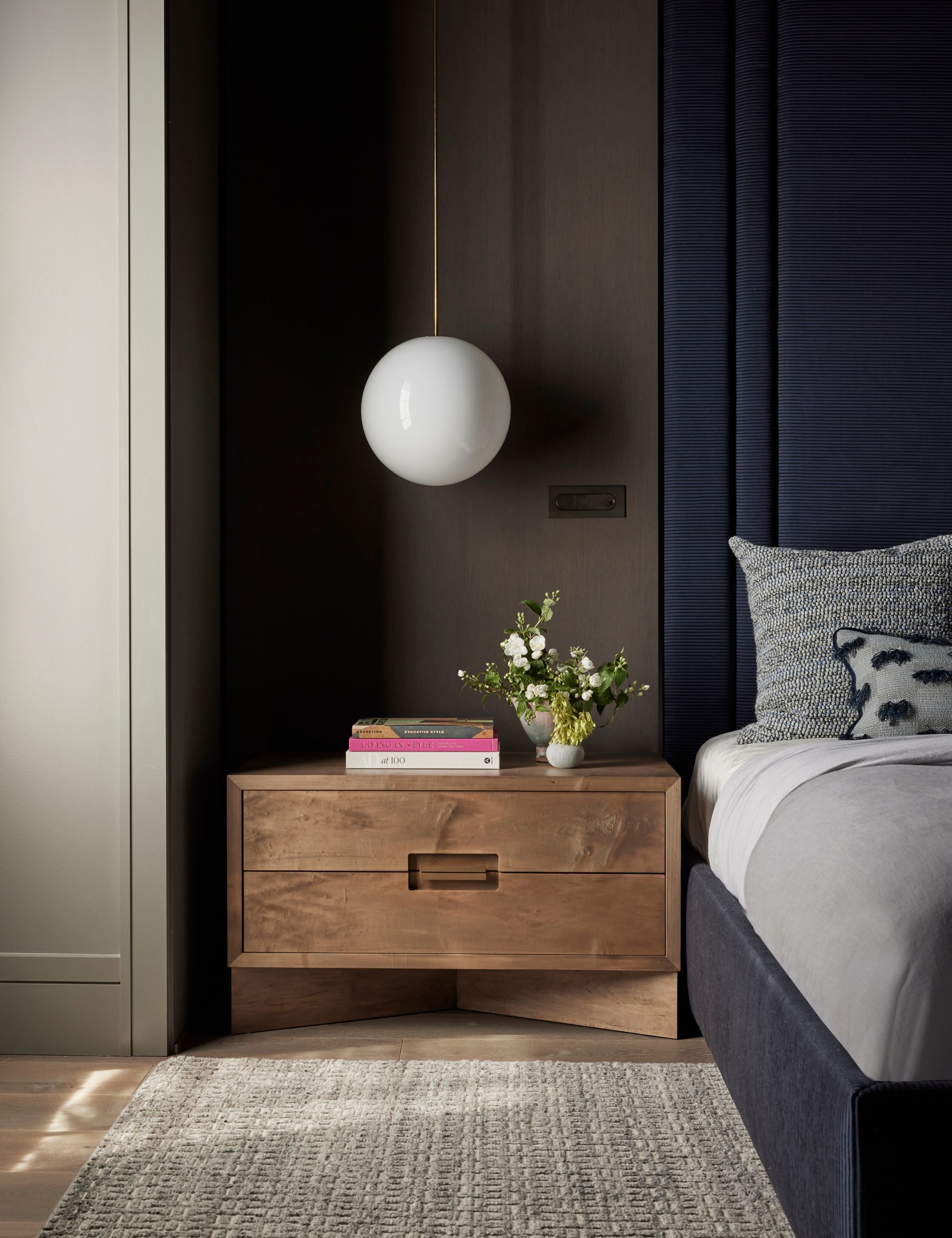 Bedroom with grey patterned rug, a white circular light hanging from the ceiling, large wooden bedside tables with books and flowers on top next to a large navy headboard with grey cushions