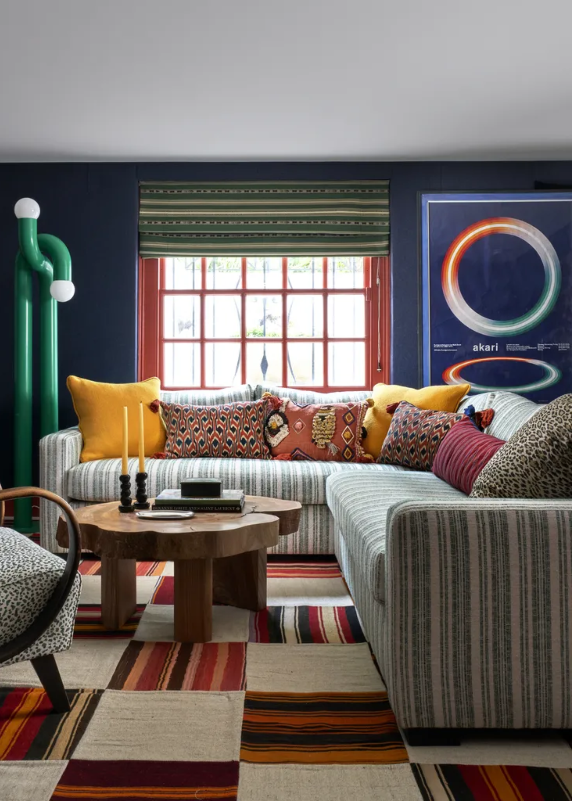 A blue living room with a striped couch and many colorful patterned pillows, a striped checkerboard rug and a wooden coffee table with candlesticks and books. A large green floor lamp is beside the couch and a large print on the wall beside it