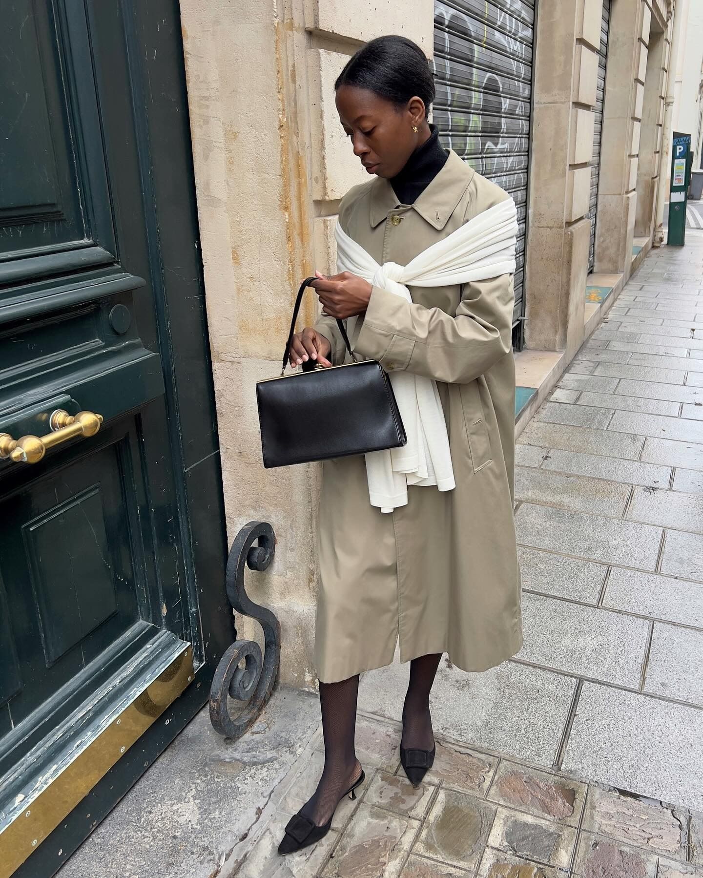 French woman wearing trench coat wrapped in white sweater, holding black bag, on the streets of Paris.