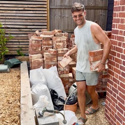 Man carrying bricks beside large pile of bricks by wooden fence in garden