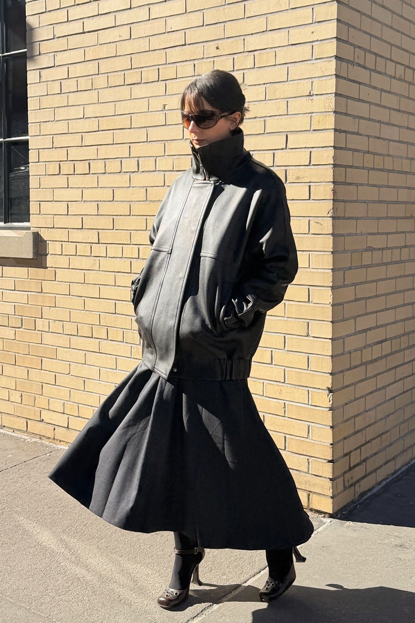 A woman wearing a black leather high collar jacket, black skirt, and heels