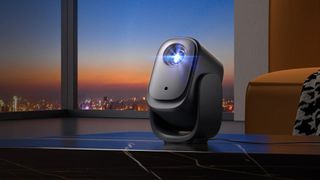 Dangbei N2 mini portable projector rendered in a lifestyle image. It's on a table pointing slightly upwards. Behind is a large window showing a city at the end of sunset.