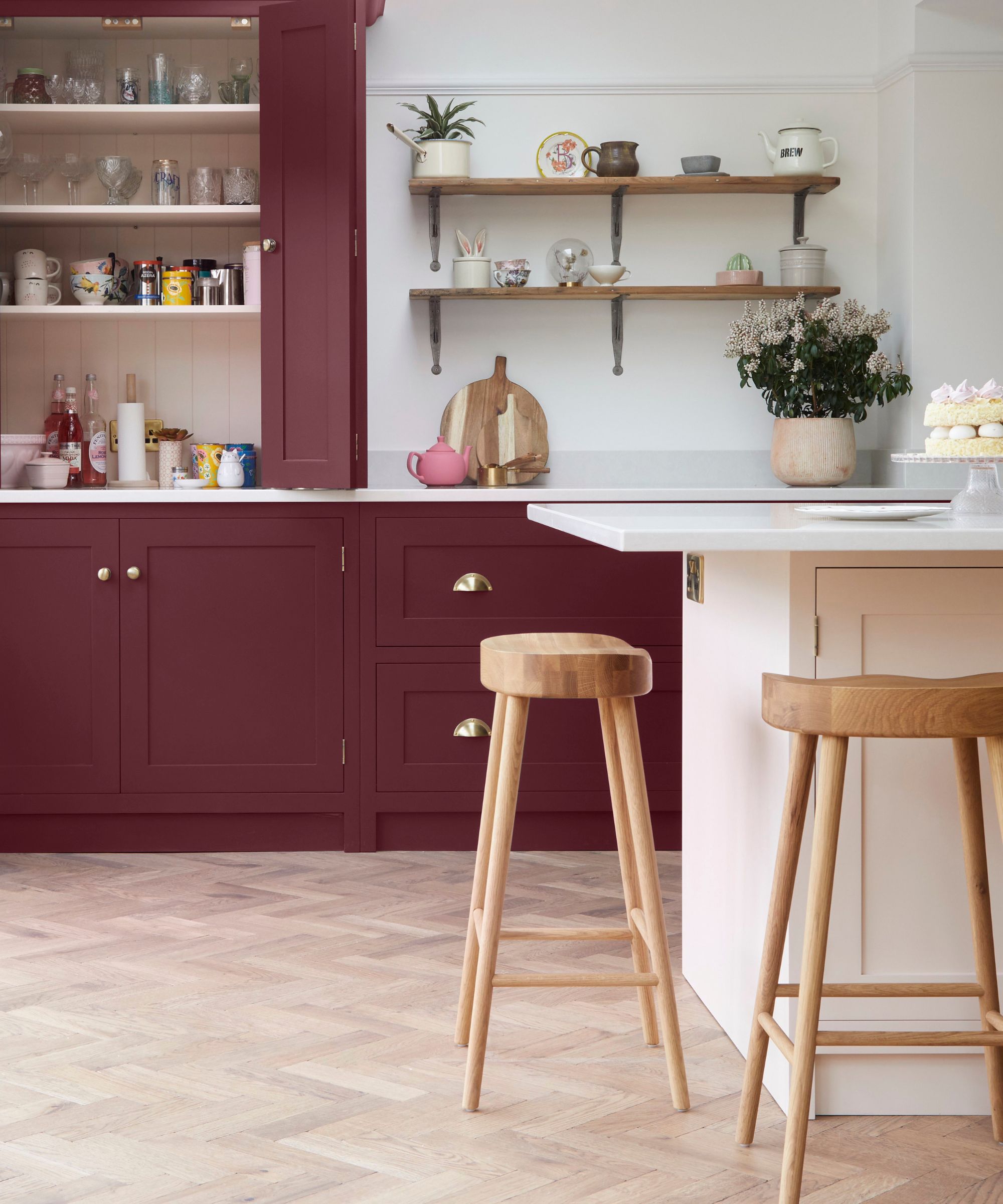 A kitchen with burgundy cabinets with open shelves with glasses and colorful mugs, wooden open shelves, and a light pink kitchen island in front with two curved wooden stools