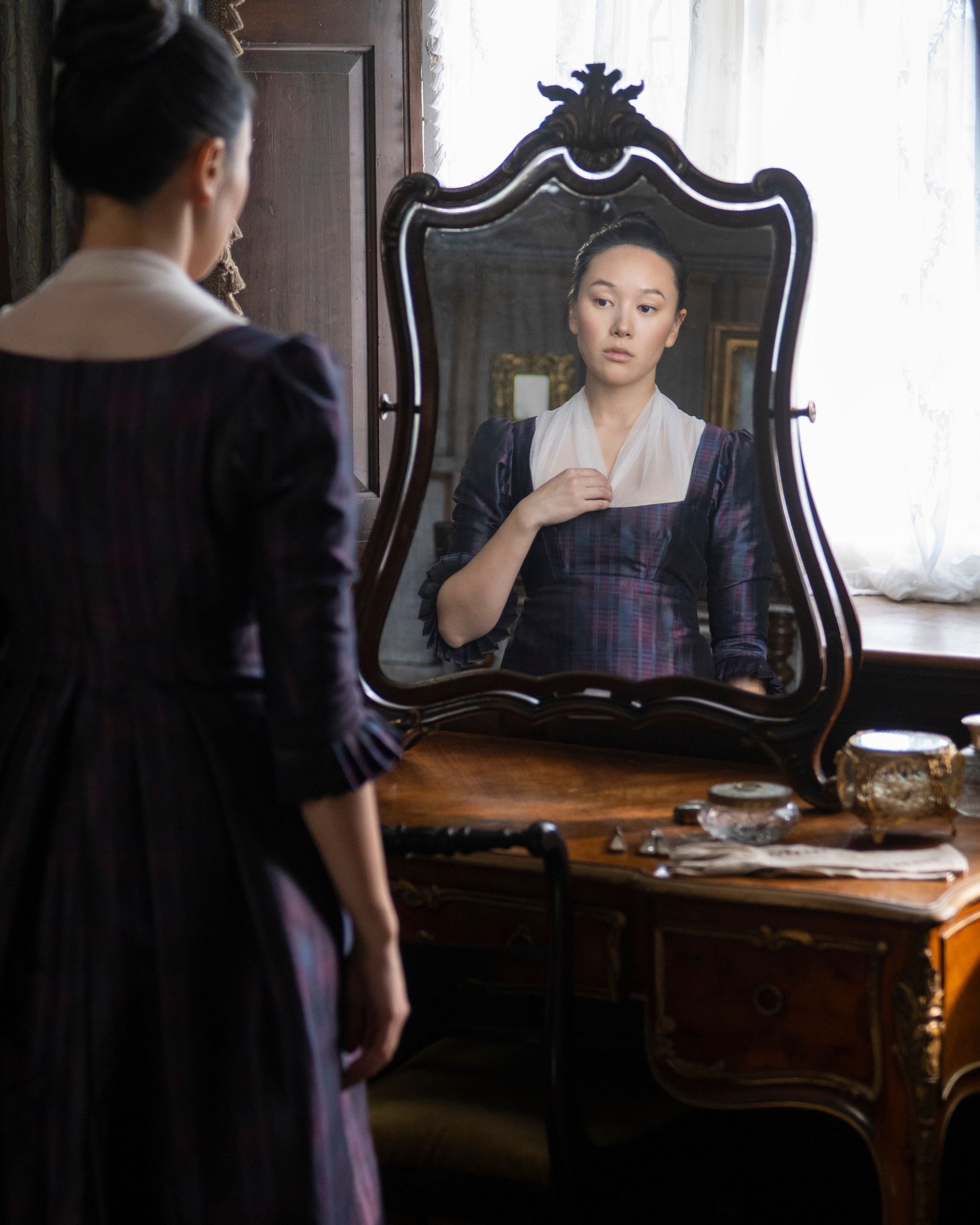 Production still from Netflix's Bridgerton series showing a woman in a dark purple gown standing, looking into an antique mirror on a wooden vanity by a window.