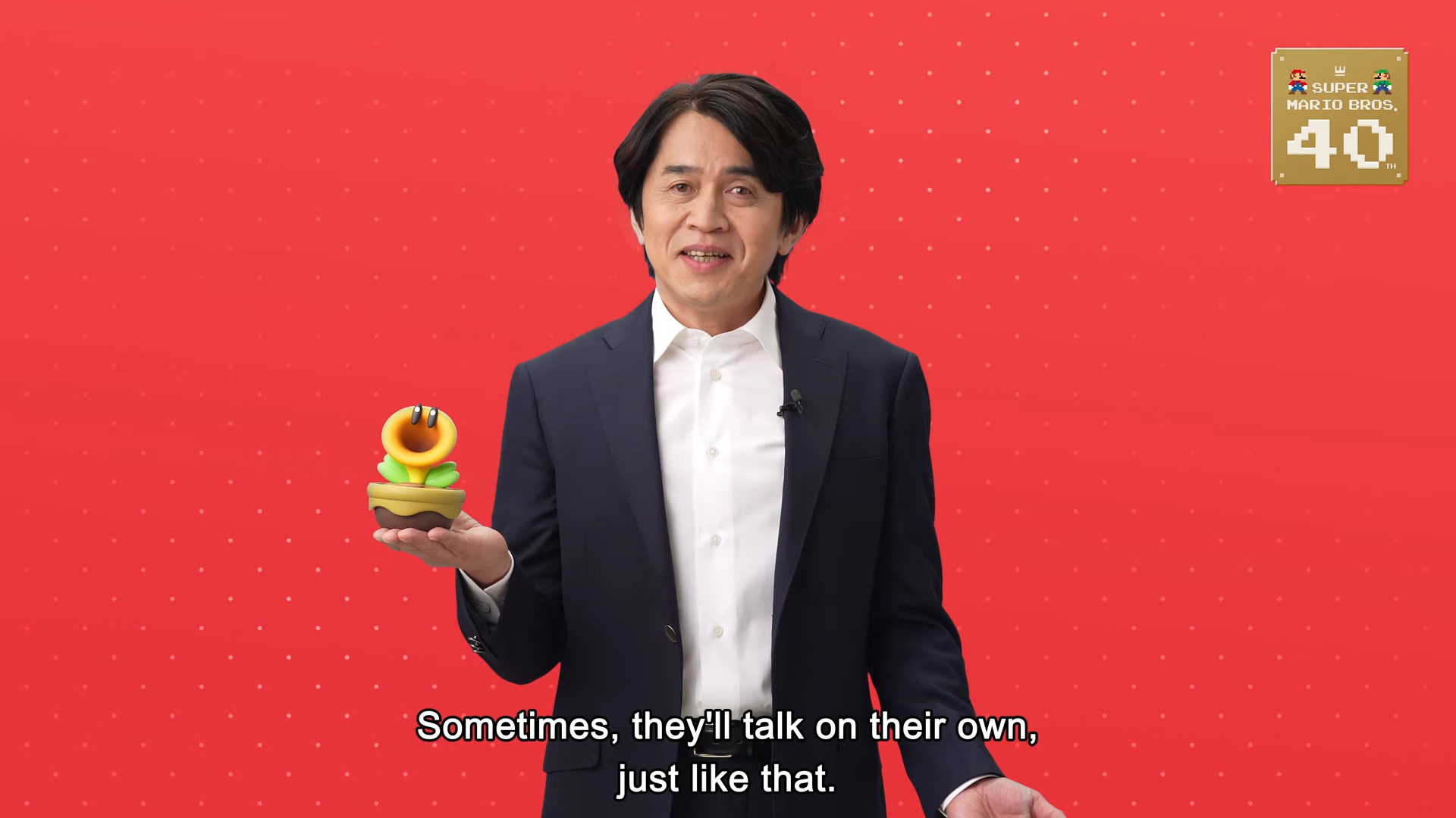 The Talking Flower being held by a man standing against a red background, with the Super Mario Bros. 40 Anniversary logo in the top right-hand corner