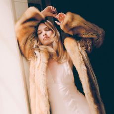 Promo image for Who What Wear's August 2025 cover story with Lily James. She is leaning up against a white post wearing a brown furry coat over a white dress with deep v neckline.