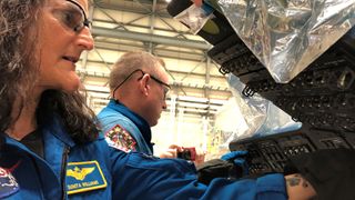 two people in blue flight suits touch control panels in an open spacecraft cockpit in a hangar