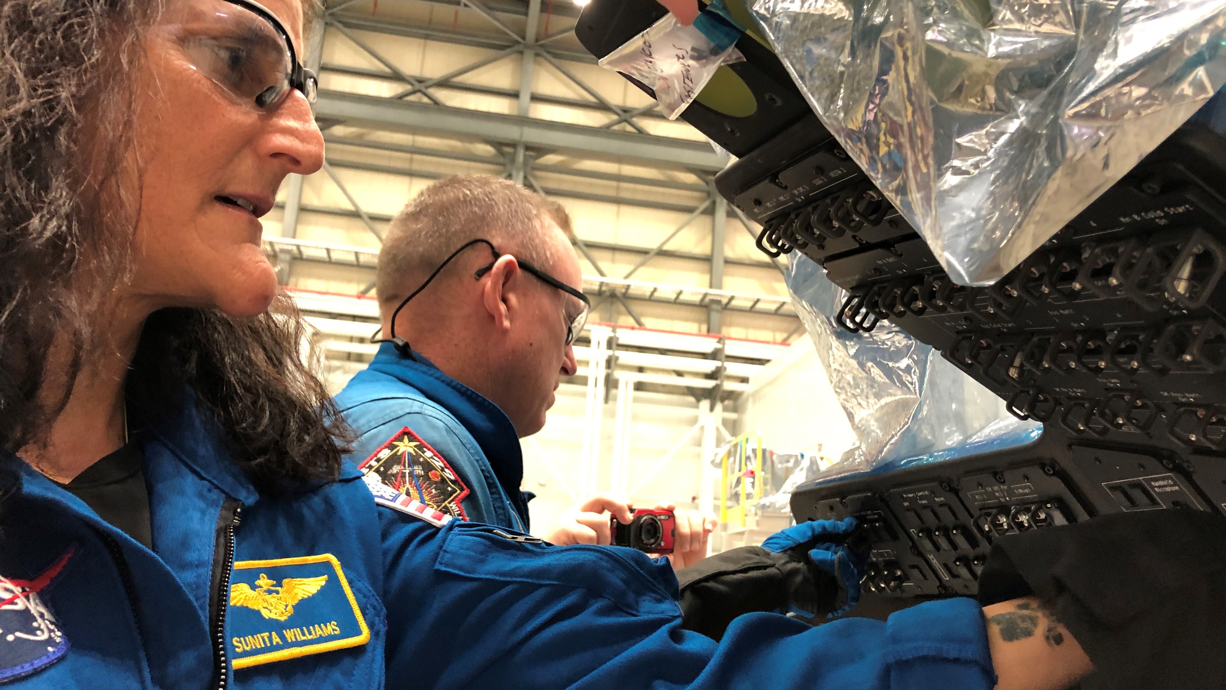 two people in blue flight suits touch control panels in an open spacecraft cockpit in a hangar