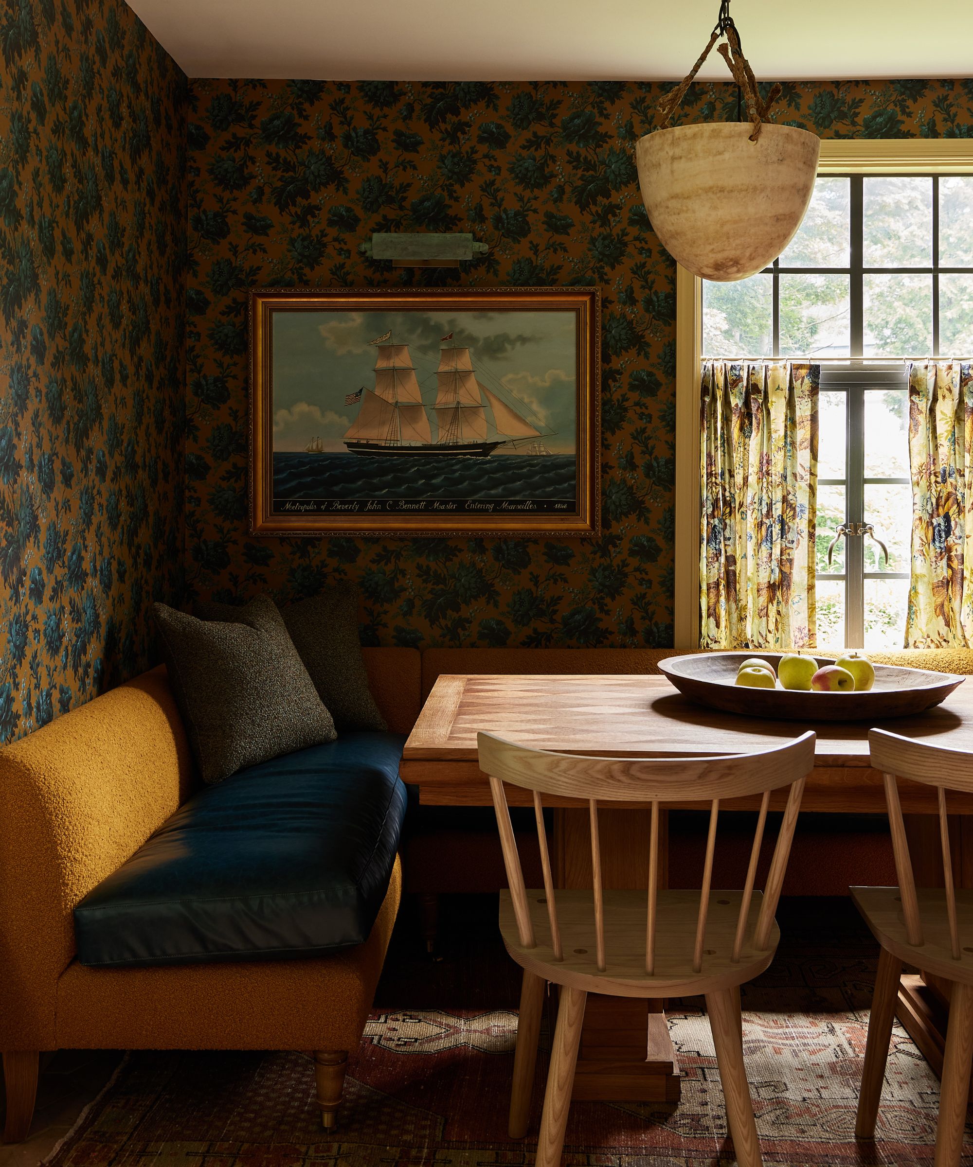 a mustard and blue floral wallpapered kitchen with a yellow banquette seat and wooden breakfast table with a seaside ship vintage painting