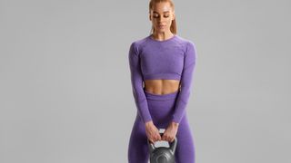 Woman in purple workout clothes holding a kettlebell in front of her with both hands against grey backdrop