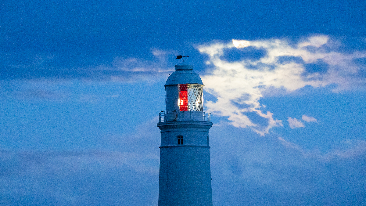 lighthouse at dusk in front of a partially covered moon