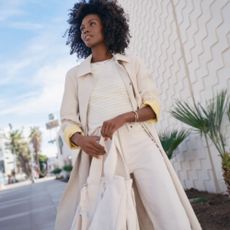 Woman wearing tailored white jeans and trench coat with yellow striped top, standing outside in street.