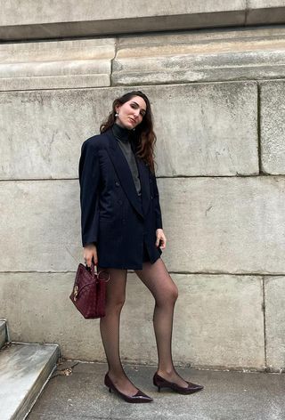 winter tight outfit shown in a photo of a woman wearing a navy blazer, gray turtleneck, black mini skirt, black fishnet tights, red kitten heels, and a red handbag