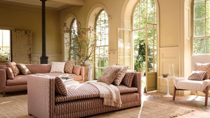 A light-filled sunroom with arched windows, pink upholstered seating, natural jute rugs, a black column, open French doors and a large flowering branch in a rustic planter.