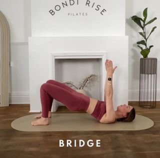 Lottie Anderson, Founder of Bondi Rise, performs a glute bridge Pilates move. She wears leggings and a matching sports bra. She is on a oval-shaped mat, in front of a decorative fireplace, performing the move. Her shoulders and feet are the only point of contact with the mat. The rest of her body is elevated in a straight line. Her arms are pointing towards the ceiling.