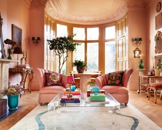 maximalist living room with pink walls, large bay window, pink armchairs and glass coffee table