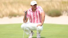 Brooks Koepka crouches down with his putter on the green at the US Open in white slacks, white cap and a pink/white polo shirt