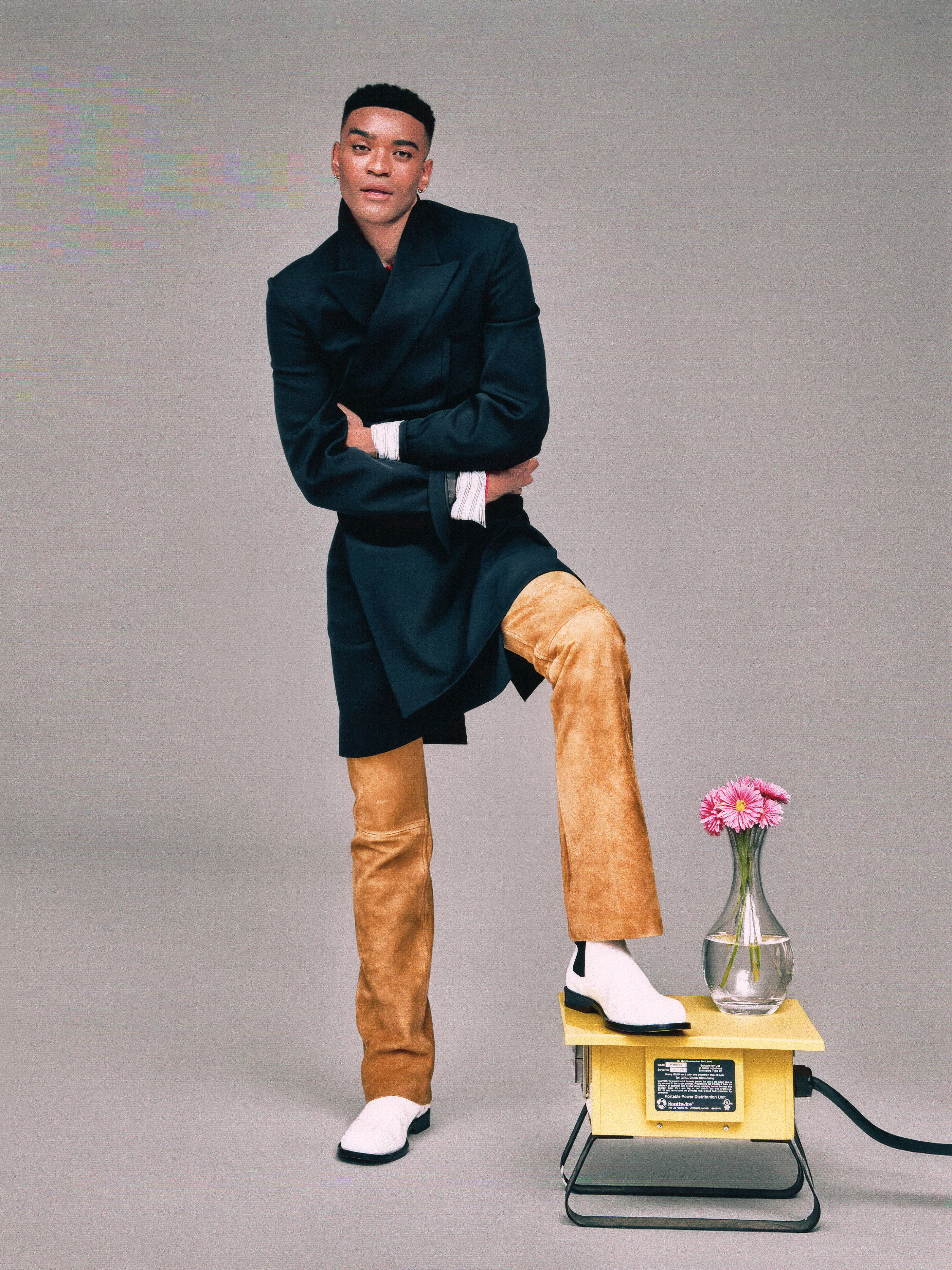 Owen Thiele poses wearing a black jacket and suede pants standing with his foot in a white boot over a vintage scale with a flower on it