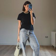 Woman wearing black t-shirt, pearl necklace, light wash jeans, and woven green bag.