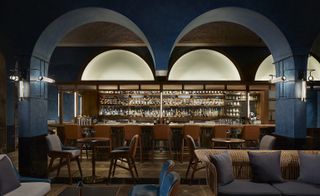 Interior view of the bar and seating area at Origin Grill & Bar, Singapore featuring dark blue walls, brown, blue and wicker seating and shelves behind the bar with multiple bottles of drinks. The room design features multiple arches