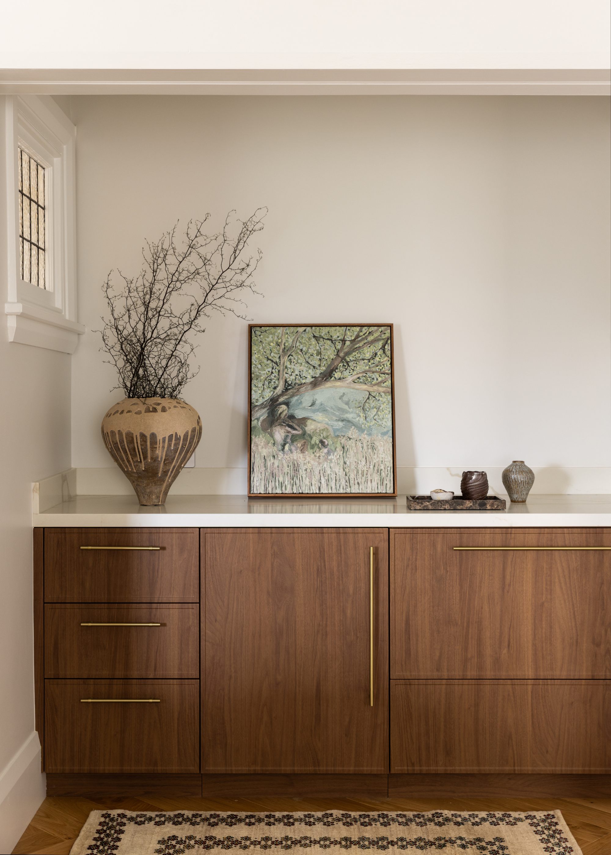 A living room with wooden cabinetry and brass hardware, a floor runner, a base with stems, framed art and mini vases