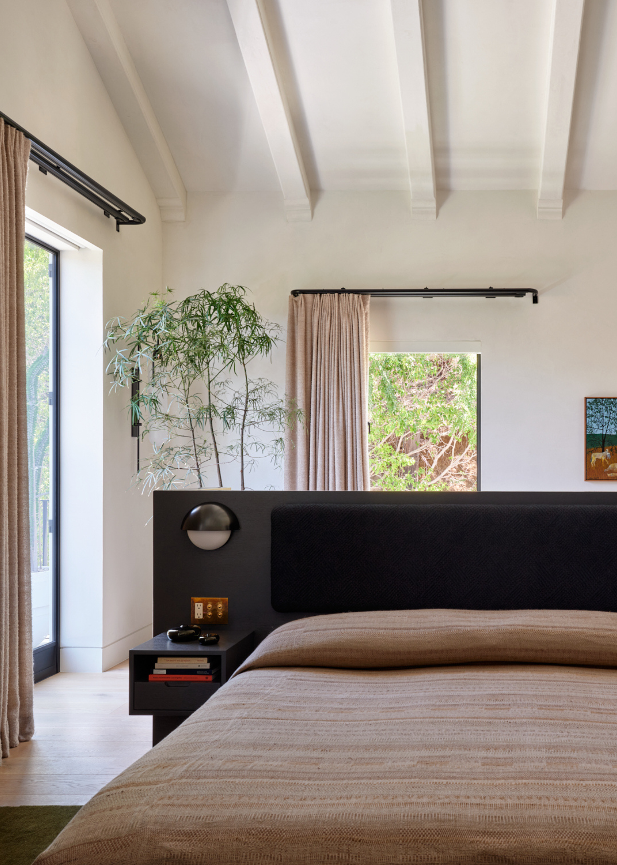 A white bedroom with vaulted ceilings, exposed beams, an extended headboard, a bed with a beige duvet and a potted tree