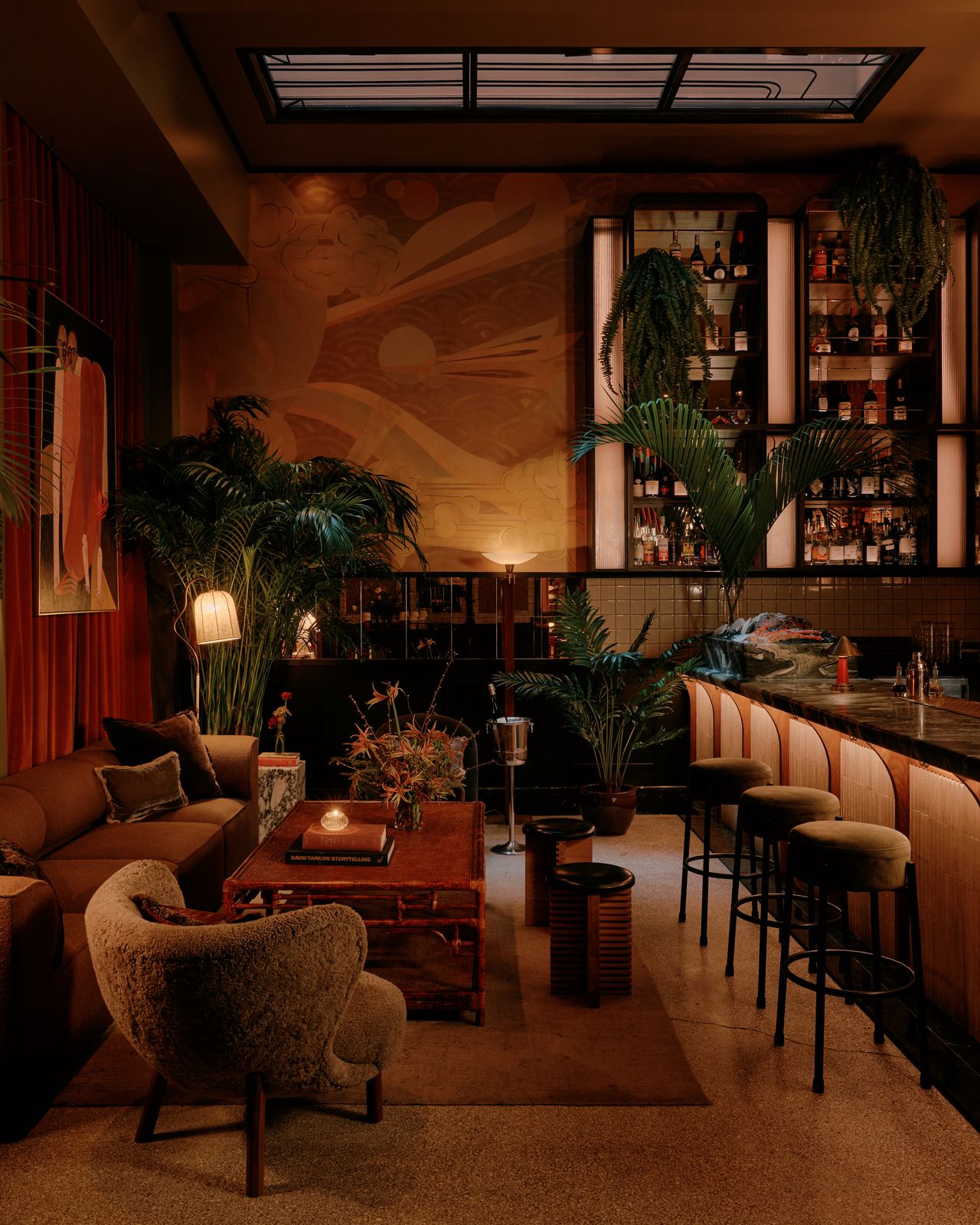 A dimly lit, imaginatively decorated cocktail bar features murals, paintings, Art Deco-inspired lighting, and sprawling plants in a warmly suffused atmosphere.