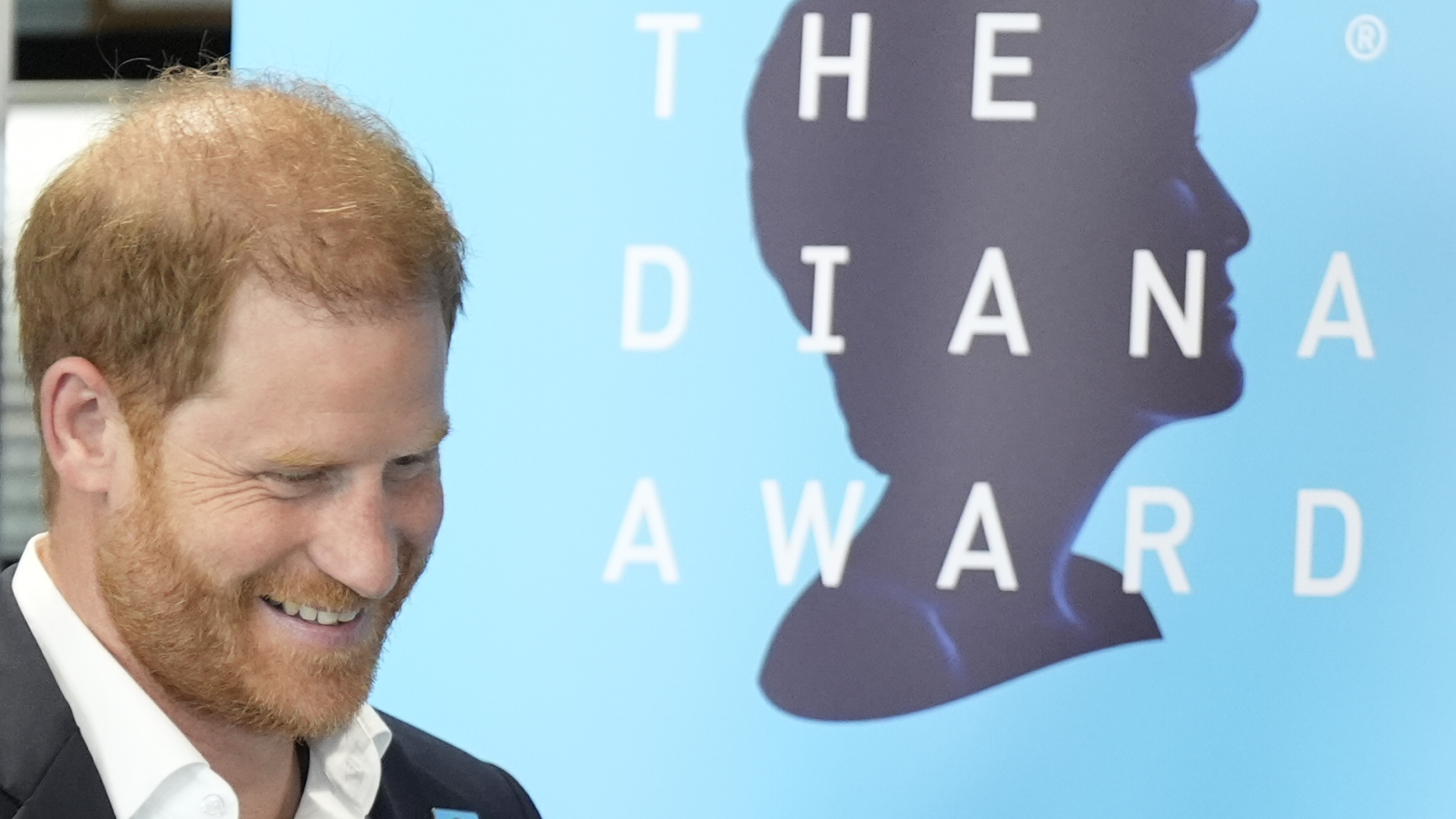 Prince Harry, in a suit and white shirt, smiling in front of a blue Diana Award backdrop