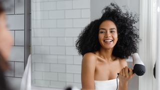 Best hair dryers 2022: A woman with curly black hair laughs while drying her hair with a white and grey blow dryer