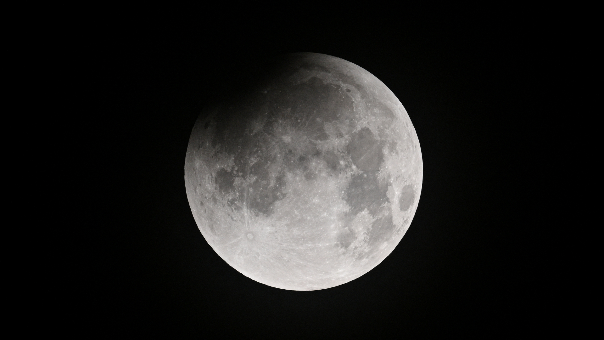 A full moon is pictured against a dark sky. Dark features called lunar maria are visible marking its surface and an impenetrable shadow has fallen over the upper right edge of the lunar disk.