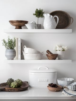 A kitchen with white shelving and accessories including potted plants, wooden pestle and mortar, bowls and jugs