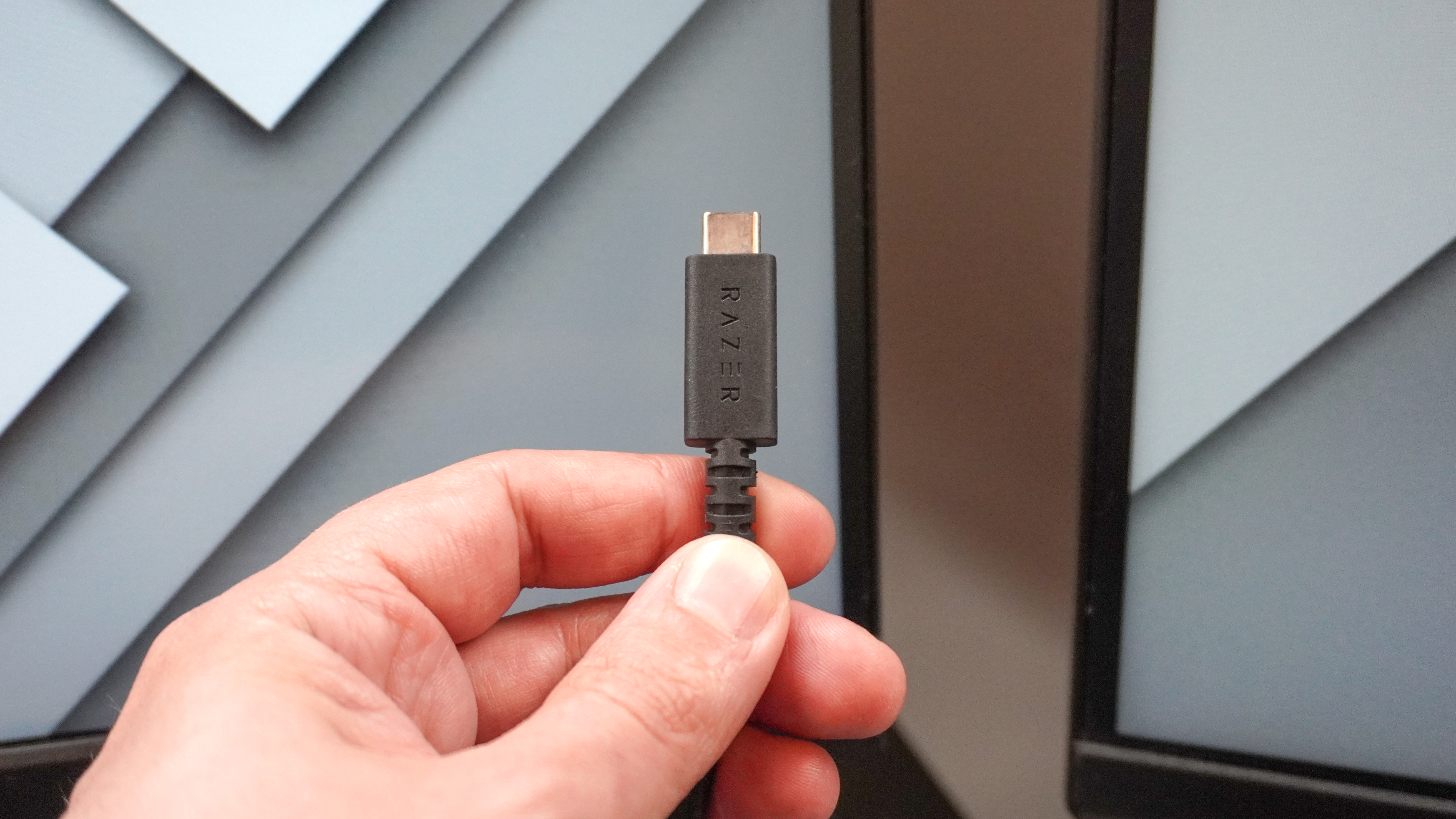 A person holding up the included USB-C to USB-C cable that comes with the Razer Kiyo V2 webcam in front of a dual monitor setup