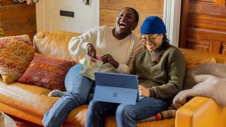 Female couple sitting on sofa with Microsoft Surface tablet, both laughing