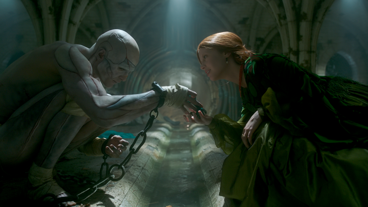 Jacob Elordi as The Creature chained up with Mia Goth holding his hand in Frankenstein