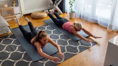 Two women lie on the floor on yoga mats performing the superman hold. They are both dressed in sportswear. They are lying on their fronts, with their arms and feet elevated. Behind them we see a cushion, a chair and a potted plant.