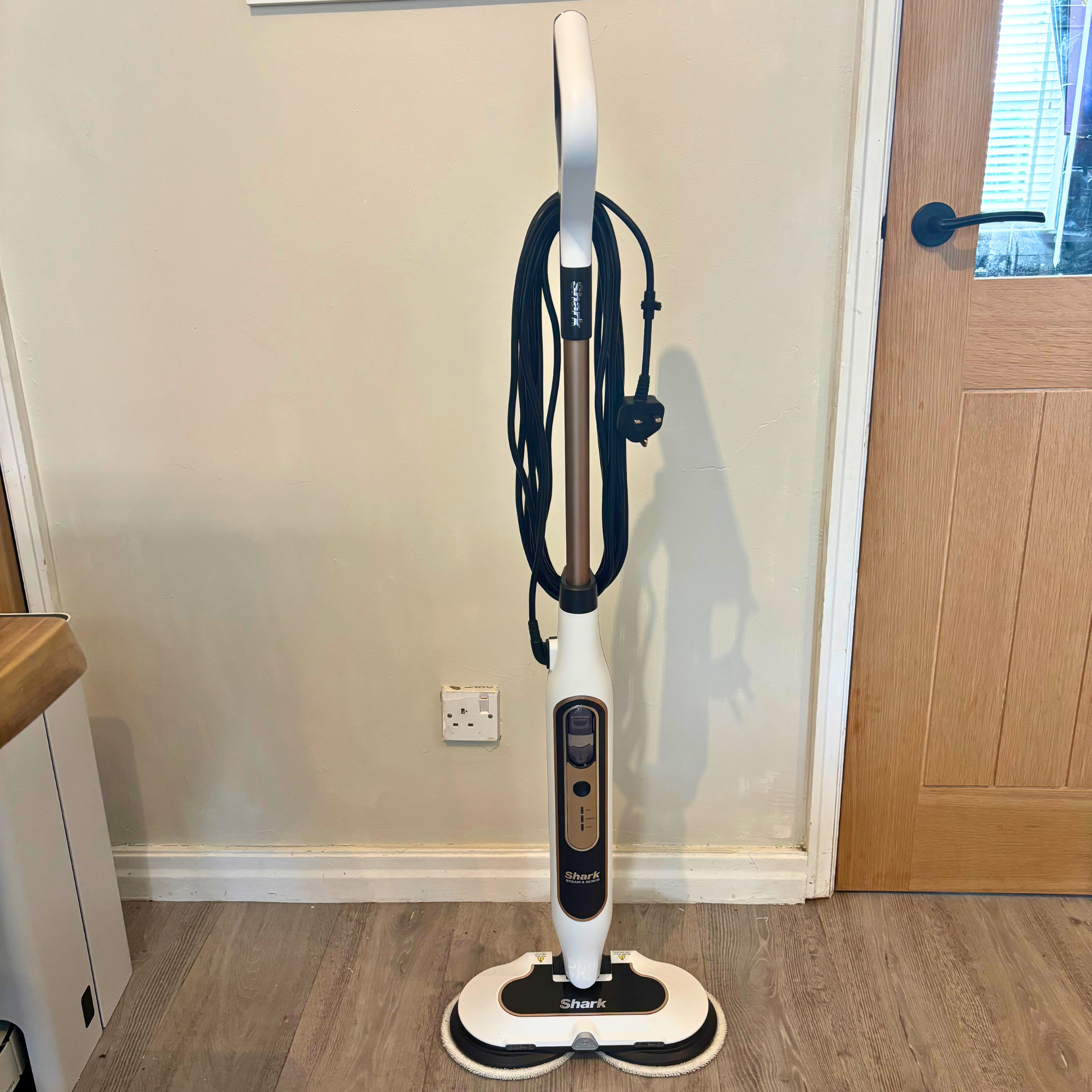 Front view of the Shark Steam & Scrub Automatic stood upright on a laminate floor in front of a cream wall and oak door