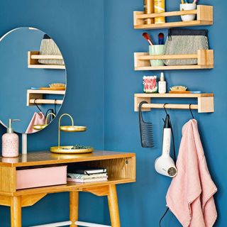 Wooden dressing table in blue painted corner with round mirror in corner beside shelves with make up and hairdryer