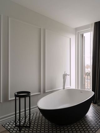 Interior view of a bathroom at The Serras featuring white walls with a rectangular panel style design, a tall window, dark curtains, a round black side table, a black and white freestanding bath, wood flooring and black and white patterned floor tiles under the bath