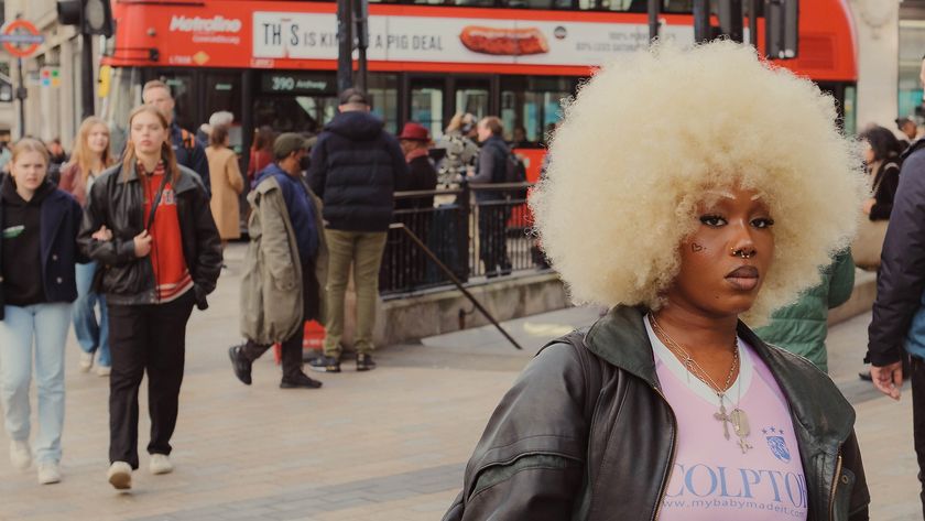 A person with a large curly afro hairstyle stands in a busy urban setting, surrounded by pedestrians and a red double-decker bus