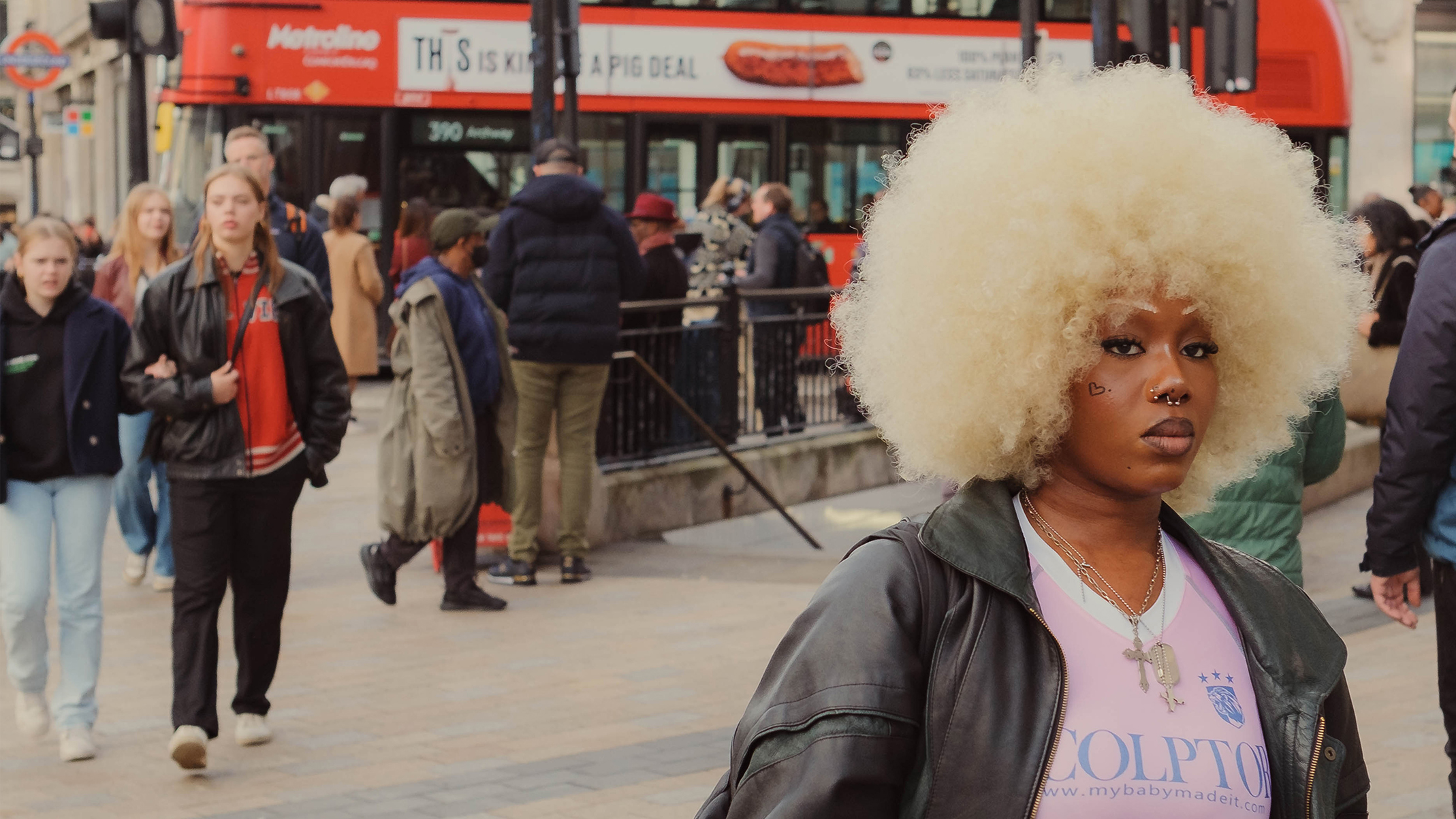A person with a large curly afro hairstyle stands in a busy urban setting, surrounded by pedestrians and a red double-decker bus.